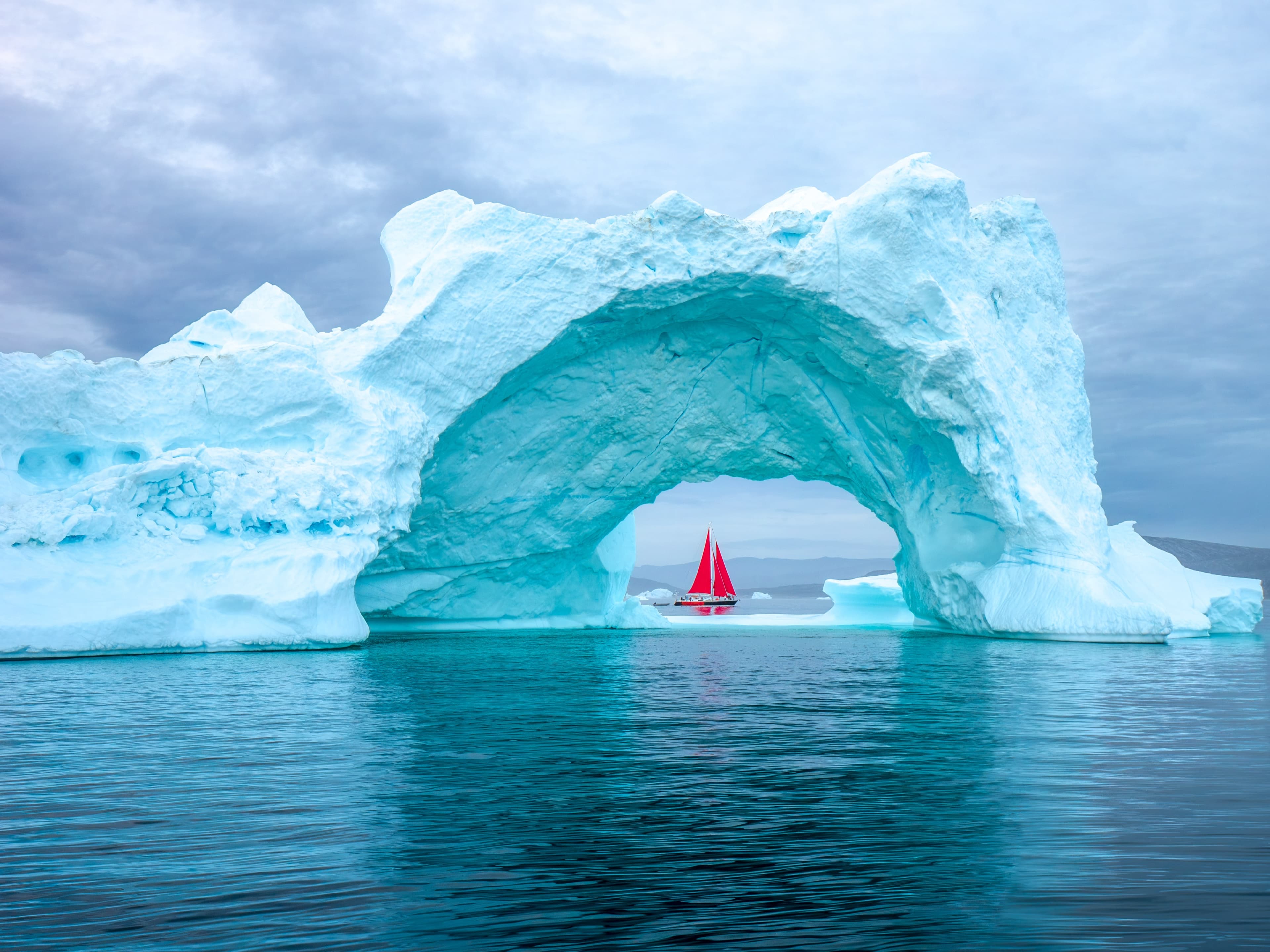 Red-sailed schooner seen through a monumental iceberg arch in Greenland — Arctic Expeditions collection